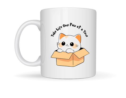“White ceramic mug featuring a cute cartoon cat in a cardboard box with the phrase ‘Take Life One Paw at a Time’ in playful script.”